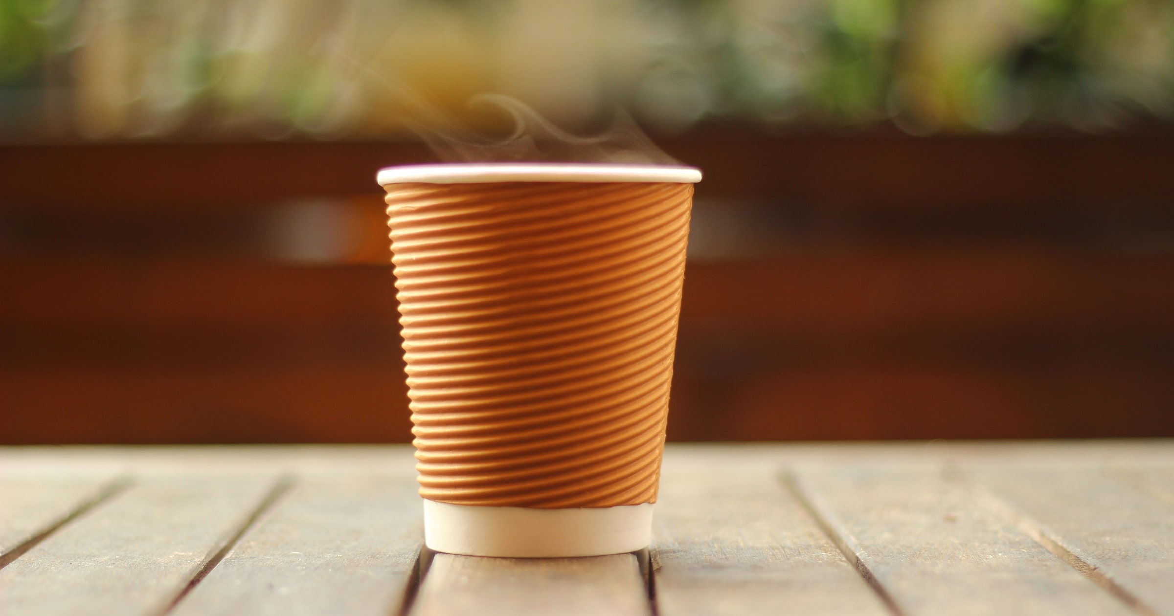 A steaming coffee in a brown paper cup on a wooden table.