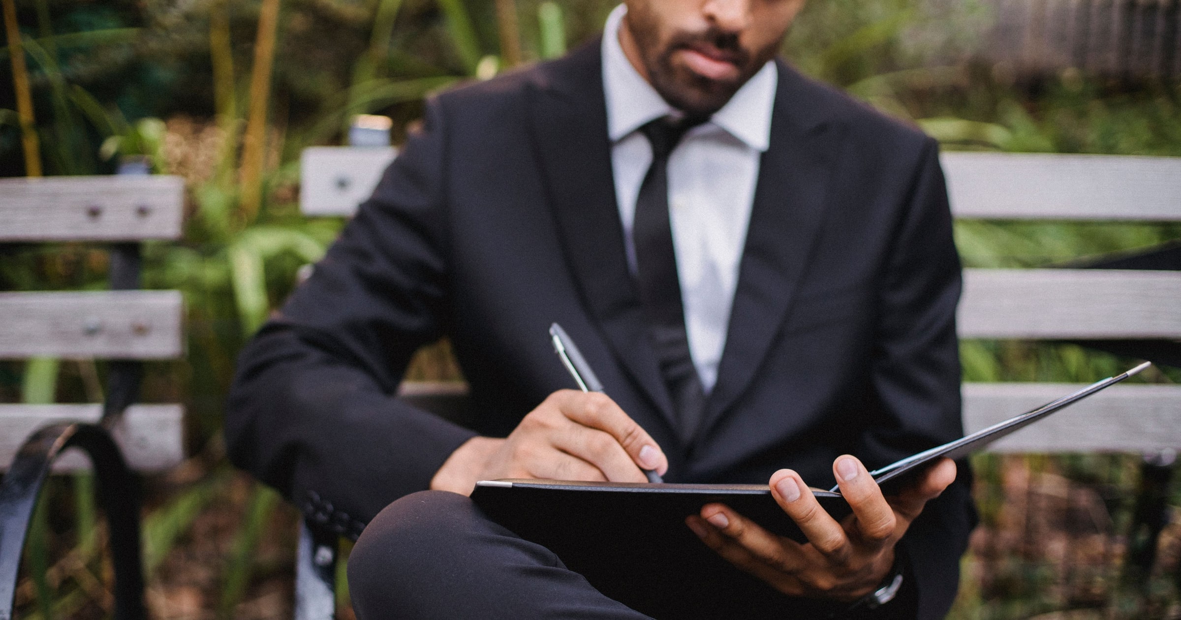 A man in a black suit writing on a clipboard while sitting on a bench.