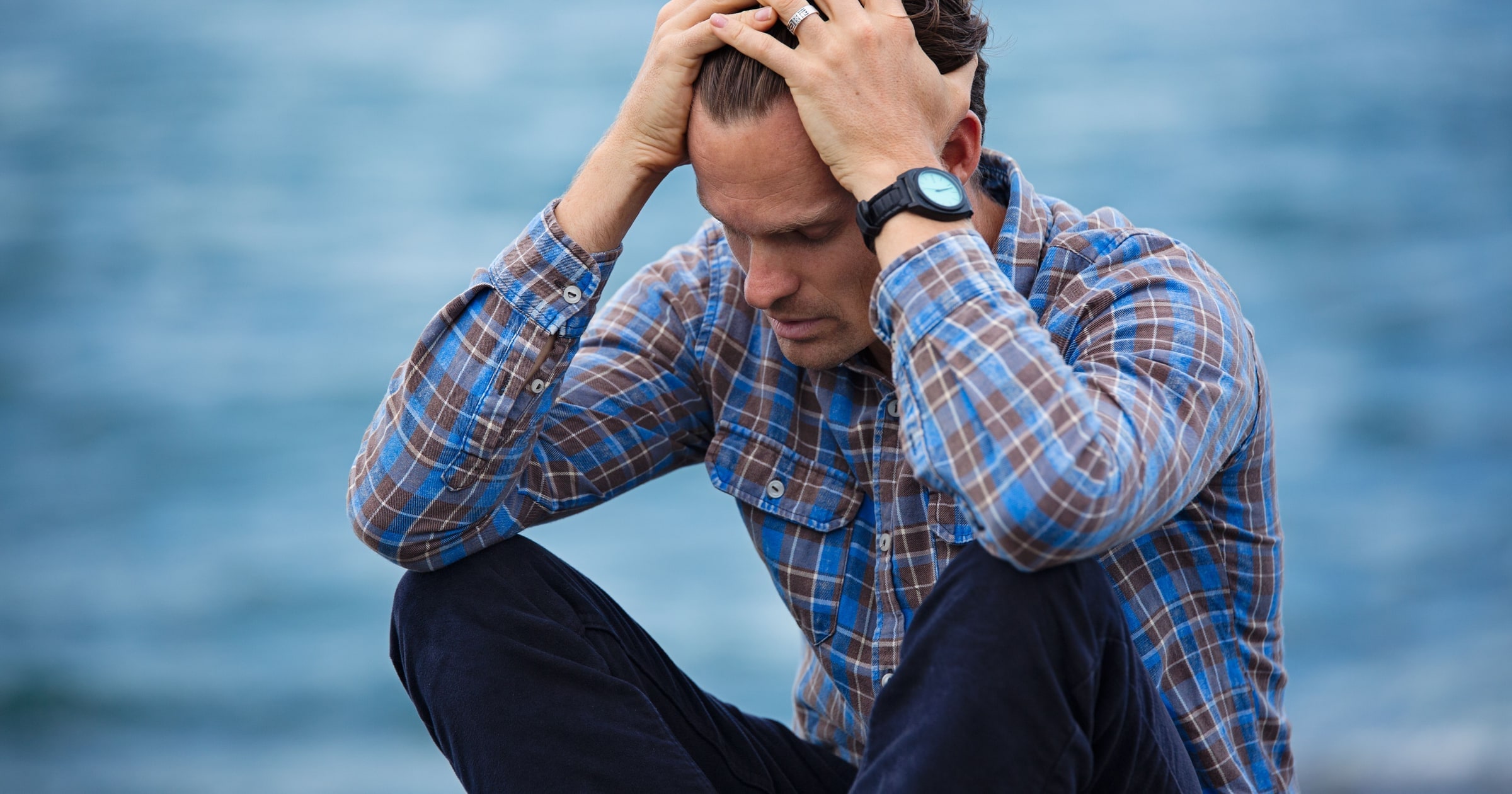 A man in a blue and brown plaid shirt sits by the water with both hands in his hair